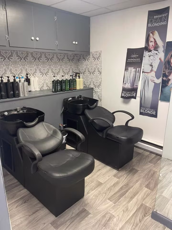 Two black shampoo bowls and chairs under gray cabinets, with a shelf of salon products and blonding posters on the wall